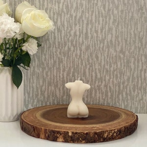 Puede incluir: Una vela blanca con forma de torso humano, sentada sobre una rodaja de madera. La vela est&aacute; sobre una superficie blanca con un arreglo floral blanco en el fondo.