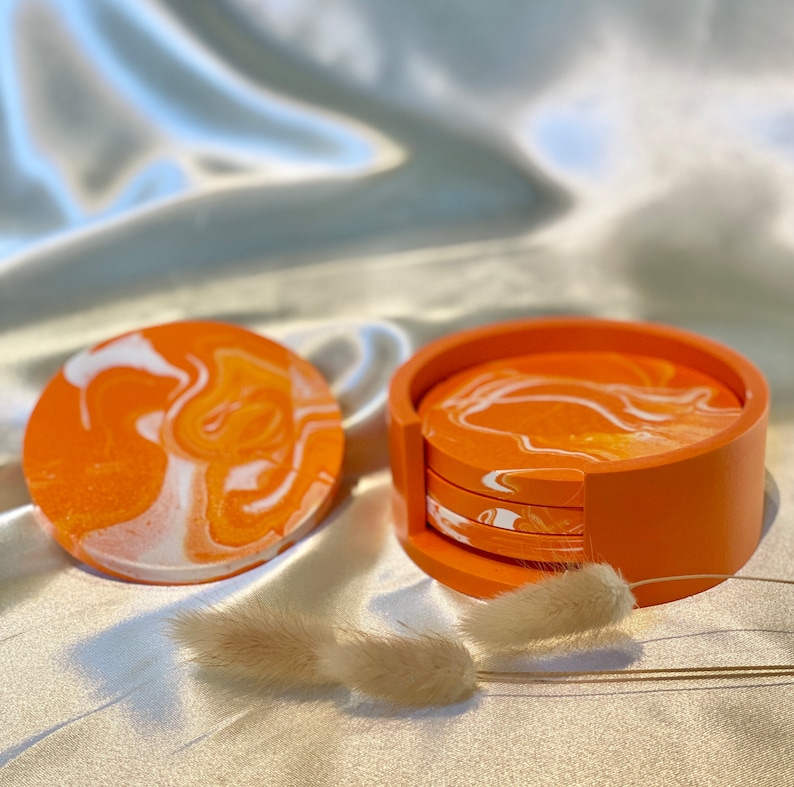 Op de afbeelding: Set van vier oranje en witte marmeren onderzetters met een bijpassende oranje houder. De onderzetters zijn rond en hebben een draaiend patroon.