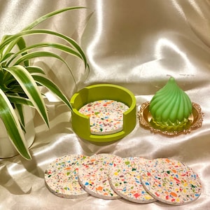 Op de afbeelding: Set van vijf kleurrijke, gevlekte onderzetters met een groene houder. De onderzetters zijn gemaakt van terrazzo en hebben een witte basis met veelkleurige vlekken. De houder is een groene ring met een uitsparing voor gemakkelijke toegang tot de onderzetters.