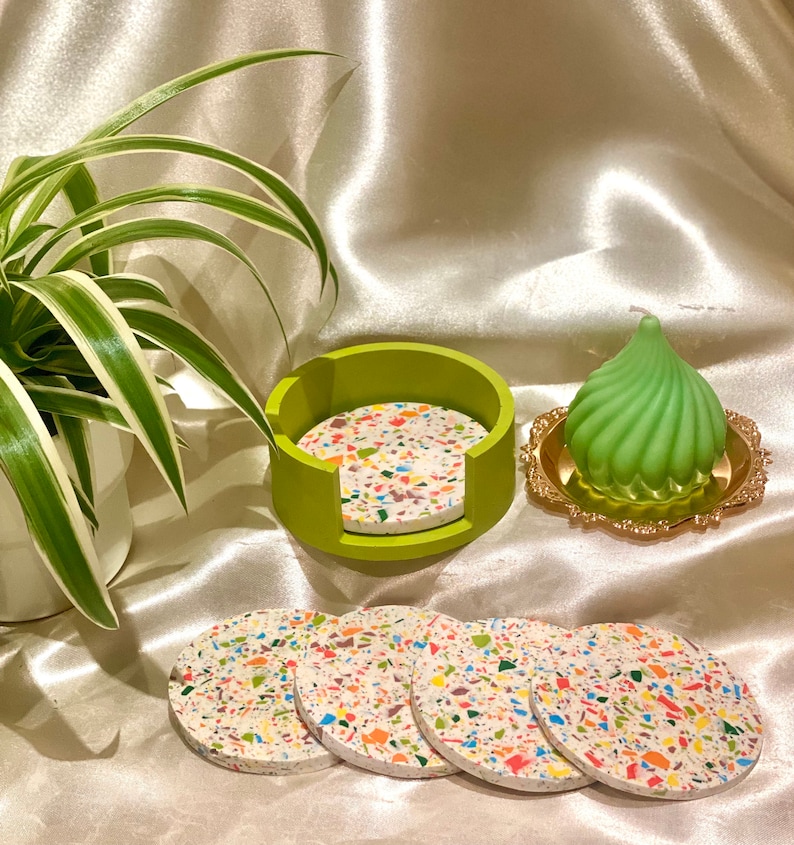 Op de afbeelding: Set van vijf kleurrijke, gevlekte onderzetters met een groene houder. De onderzetters zijn gemaakt van terrazzo en hebben een witte basis met veelkleurige vlekken. De houder is een groene ring met een uitsparing voor gemakkelijke toegang tot de onderzetters.