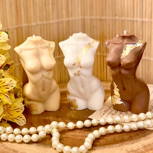 May include: Three decorative candles shaped like the human torso, each with a different skin tone. The candles are decorated with gold leaf accents. The candles are sitting on a wooden surface with a string of pearls in front of them.