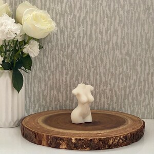 Puede incluir: Una vela blanca con forma de torso humano, sentada sobre una rodaja de madera. La vela est&aacute; sobre una superficie blanca.