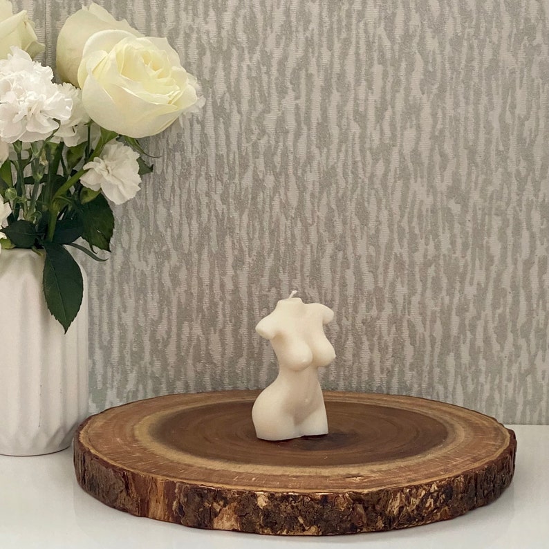 Puede incluir: Una vela blanca con forma de torso humano, sentada sobre una rodaja de madera. La vela est&aacute; sobre una superficie blanca.