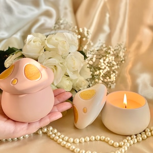 Op de afbeelding: Twee keramische kaarshouders in de vorm van een paddenstoel met gouden accenten. De ene houder is roze en de andere is wit. De witte houder heeft een kaars met een brandende lont. De kaarshouders staan op een oppervlak met witte parels.