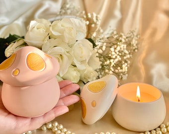 May include: Two ceramic mushroom-shaped candle holders with gold accents. One holder is pink and the other is white. The white holder has a candle inside with a lit wick. The candle holders are on a surface with white pearls.