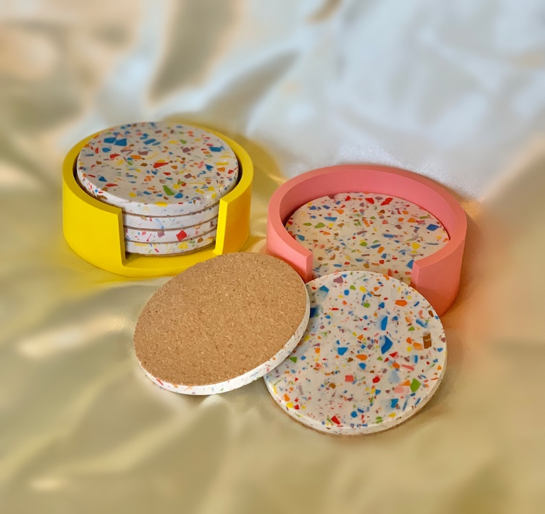 Op de afbeelding: Set van zes kleurrijke terrazzo-onderzetters in een gele houder en een set van vier terrazzo-onderzetters in een roze houder. De onderzetters zijn gemaakt van een witte basis met kleurrijke vlekken van terrazzo.