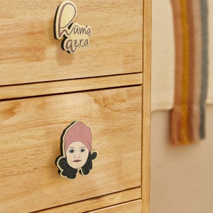 Könnte beinhalten: Eine Kommode aus Holz mit zwei Schubladen. Die obere Schublade hat einen Holzgriff mit dem Text "Wüma azra" darauf. Die untere Schublade hat einen Holzgriff mit einem Cartoon-Bild einer Person mit einem rosa Hut.