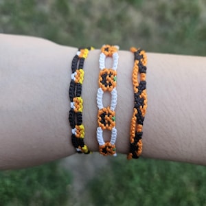 May include: Three Halloween-themed friendship bracelets. One has a candy corn design in black, yellow, and white. Another features orange pumpkin shapes with green details. The third bracelet has an orange and black pattern.