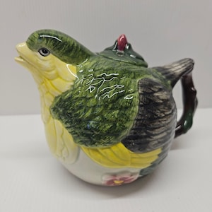 May include: A ceramic teapot shaped like a bird, featuring a yellow beak and chest, green wings, and a gray tail. The teapot has a small red accent on the lid and a brown handle. The bird-shaped teapot is a unique kitchenware item.