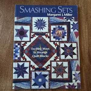 Op de afbeelding: Een boek getiteld "Smashing Sets" van Margaret J. Miller. De omslag toont een patchwork-ontwerp met ster- en vierkante quiltblokken in tinten blauw, paars en rood. De tekst "Exciting Ways to Arrange Quilt Blocks" is ook zichtbaar.