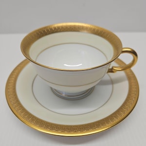 May include: A white teacup and saucer set with gold trim. The teacup has a gold handle and a gold band around the rim. The saucer has a matching gold border. The set is likely made of porcelain.