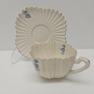 May include: A cream-coloured teacup and saucer set. The cup and saucer have a scalloped edge with gold trim. Blue floral accents are placed on the cup and saucer. The cup has a unique handle design.