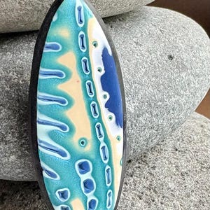 May include: An oval-shaped pendant with a black border and a colorful, abstract design. The design features shades of blue, teal, white, and yellow, creating a unique and eye-catching pattern. The pendant is likely made of polymer clay.
