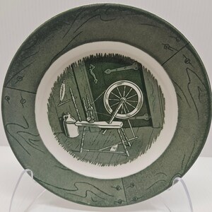 Può includere: Un piatto rotondo in ceramica con un bordo verde scuro e un motivo a linee. Il centro mostra un'illustrazione in bianco e nero di un filatoio, una brocca e una porta in legno con cerniere in metallo, il tutto in uno stile a schizzo.