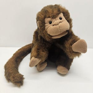 Può includere: Una scimmia di peluche con pelliccia marrone e viso, mani e piedi color beige. La scimmia ha una lunga coda ed è seduta su una superficie bianca. Il giocattolo ha occhi neri e un'espressione amichevole.