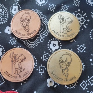 May include: Four round metal tokens with a cartoon cherub design. Each token features a different cherub, one with angel wings, one with devil horns, and two with haloes. The tokens are engraved with the text "Helluva Hanks".