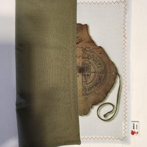 May include: An olive green fabric sleeve with gold stitching partially covers a white fabric with a vintage map design. The map has a brown, aged appearance with text and a seal. A small tag with text and a red apple is visible.