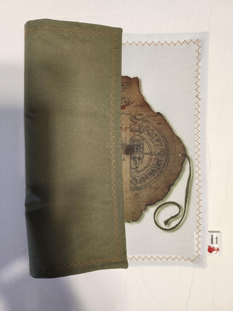 May include: An olive green fabric sleeve with gold stitching partially covers a white fabric with a vintage map design. The map has a brown, aged appearance with text and a seal. A small tag with text and a red apple is visible.
