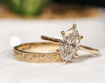 Peut inclure: Deux bagues en or. L'une présente un diamant taille marquise. L'autre a un motif gravé et orné. Les bagues sont posées sur un fond blanc doux, avec une plante verte floue en arrière-plan.