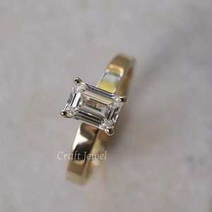 Może przedstawiać: Złoty pierścionek z przezroczystym szmaragdowym kamieniem szlachetnym. Pierścionek znajduje się na białym tle. Craft Jewel.