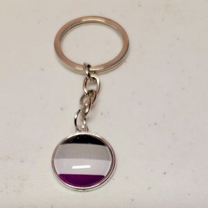May include: A silver keychain with a round, glass charm featuring the asexual pride flag. The flag has black, white, gray, and purple stripes.