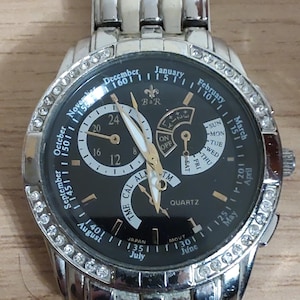 Op de afbeelding: Een zilveren polshorloge met een zwarte wijzerplaat en een bezel bezet met kristallen. De wijzerplaat van het horloge heeft een datumaanduiding, een dag van de week-aanduiding en een 24-uurs tijdweergave. De wijzerplaat van het horloge bevat ook de tekst "TIME CAL ALA TM" en "QUARTZ".