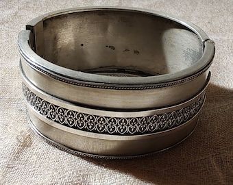 May include: A silver bangle bracelet with an intricate design. The bracelet is made of a smooth metal and has a polished finish.