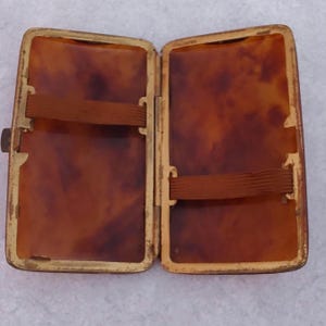 Puede incluir: Estuche de cigarrillos vintage abierto con un patrón de carey. El estuche es rectangular con un marco de metal dorado y dos bandas elásticas marrones en el interior. El estuche está abierto, revelando el interior.
