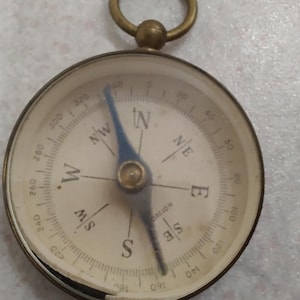 May include: A vintage brass compass with a clear glass face. The compass needle points north. The dial features directional markings and degree indicators. A small brass ring is attached at the top.