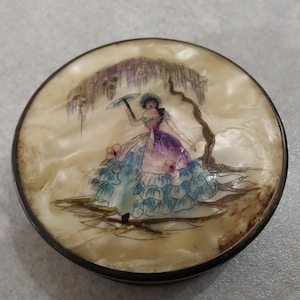 Puede incluir: Una caja redonda de color crema para guardar pequeños objetos con un borde de metal. La tapa presenta una escena pintada de una mujer con un vestido azul y blanco que sostiene un paraguas, de pie bajo un árbol.