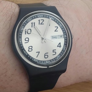 Puede incluir: Un reloj de pulsera negro con una esfera blanca y agujas plateadas. La esfera del reloj tiene números negros y una pequeña ventana de fecha que indica "THU 14".