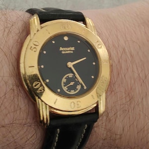 May include: A gold-tone wristwatch with a black face and a black leather band. The watch face has gold-tone hour markers and hands, and a small sub-dial at the 6 o'clock position. The watch face also features the text "Accurist Quartz".