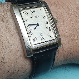 Puede incluir: Un reloj Rotary rectangular con caja plateada y esfera blanca con números romanos. Tiene una correa de cuero negro. El reloj está en una muñeca.