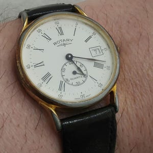 Peut inclure: Une montre-bracelet Rotary quartz en or avec un cadran blanc et un bracelet en cuir noir. Le cadran de la montre comporte des index en chiffres romains et une fenêtre de date à 3 heures. Le cadran de la montre comporte également un petit cadran avec les mots "40 Quartz" imprimés dessus.