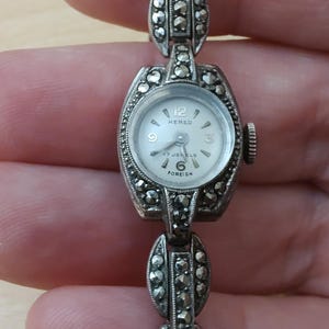 May include: A silver watch with a white face and black markings. The watch has a silver band with marcasite stones. The watch face says "Kered" and "17 Jewels Foreign".