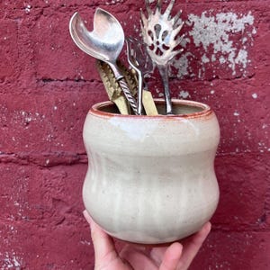 May include: A cream-colored ceramic utensil holder with a red rim, holding various silver and gold kitchen tools. The holder has a rounded shape and is held in a hand against a red brick wall.