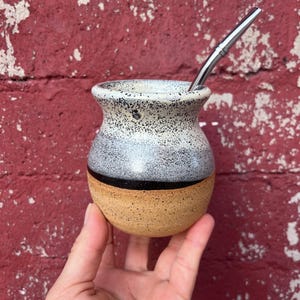 May include: A ceramic mate gourd featuring a speckled white and grey upper section, a solid black horizontal band, and a natural tan speckled base. A slender, polished stainless steel bombilla straw is inserted into the top of the rounded vessel.