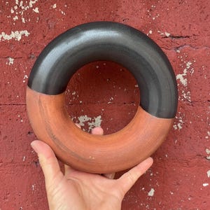 May include: A ceramic ring with a two-tone design. The top half is a dark gray, and the bottom half is a terracotta color. The ring is held in a hand against a red brick wall.