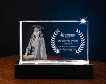 Könnte beinhalten: Ein klarer Glas-Abschlussblock mit einem Foto einer Absolventin in Hut und Robe. Der Block zeigt den Text "De Montfort University Leicester", den Namen der Absolventin und die Abschlussinformationen. Er steht auf einem schwarzen Sockel.