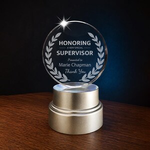 May include: A clear, round crystal award with the text "HONORING A VERY SPECIAL SUPERVISOR" and a laurel wreath design. The award is presented to Marie Chapman and says "Thank You". It sits on a silver-colored base.