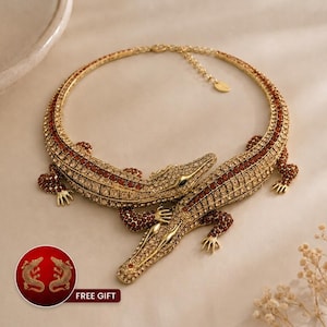 May include: A gold-coloured necklace with two crocodile figures, adorned with clear and red crystals. The necklace has a circular design and a chain clasp. The image also shows a small graphic with two crocodile earrings and the text "FREE GIFT".