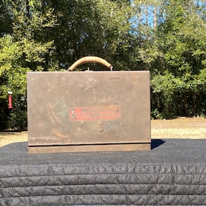 Peut inclure: Une boîte à outils en métal vintage avec une poignée tressée. La boîte à outils est rectangulaire, avec une finition marron vieillie et une étiquette rouge et blanche qui dit "Mossberg". La boîte à outils est posée sur une surface noire.