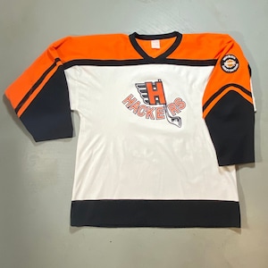 Peut inclure: Maillot de hockey blanc avec des accents orange et noirs. Le maillot présente le mot "HACKERS" en orange, avec un logo "H" stylisé et une crosse de hockey. Un écusson sur la manche indique "HIGH BLOCK HARLEY-DAVIDSON".