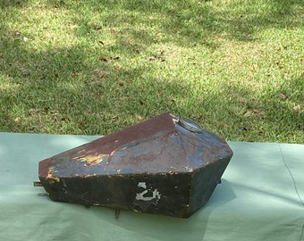 May include: A vintage, geometric-shaped motorcycle gas tank with a rusty, weathered appearance. The tank is primarily dark gray with areas of rust and yellow paint. It has a circular fuel cap and is set on a green surface.