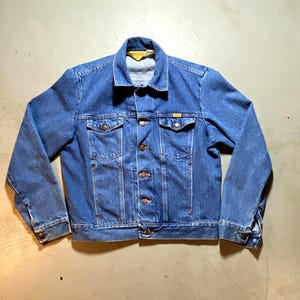 May include: A medium-wash blue denim jacket featuring a pointed collar, long sleeves with buttoned cuffs, and a front button closure. The jacket includes two chest pockets with buttoned flaps and a yellow brand label inside the collar.