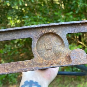 Puede incluir: Primer plano de una conexi&oacute;n de tuber&iacute;a de metal oxidado con dos agujeros ovalados y una abertura circular con el texto "MADE IN USA" en su interior. La conexi&oacute;n es probablemente para un sistema de plomer&iacute;a o de riego.