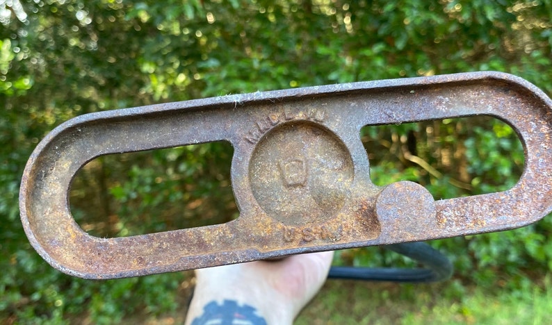 Puede incluir: Primer plano de una conexi&oacute;n de tuber&iacute;a de metal oxidado con dos agujeros ovalados y una abertura circular con el texto "MADE IN USA" en su interior. La conexi&oacute;n es probablemente para un sistema de plomer&iacute;a o de riego.