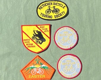 May include: Five embroidered patches featuring bicycle-themed designs. The patches are yellow, orange, and white with black and red accents. The patches feature text that reads "Metuchen Bicycle Touring Society", "Bike Tour 1975", "Swiss Days Berne Indiana", "Big Law Easter", and "Nassau County Council 1962 Bike-A-Thon BSA Merroque Dist".