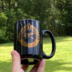 May include: A black ceramic mug featuring a gold-colored circular emblem with a clock face design. The text reads Simplex, 1894, 1994, and 100 YEARS. The emblem is surrounded by a laurel wreath and a star at the top, centered on the front of the mug.
