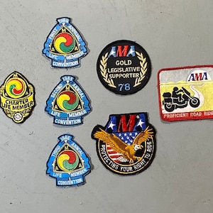 May include: A collection of vintage motorcycle patches featuring the AMA logo. Patches include "Charter Life Member", "Gold Legislative Supporter", and convention badges from 1984, 1985, and 1986. The patches are in various shapes and colors.